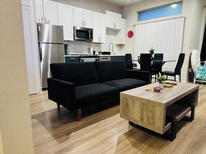 Un espace de vie moderne avec un canapé noir, une table basse en bois et un coin cuisine doté de meubles blancs et d’appareils électroménagers en acier inoxydable.