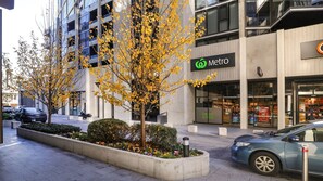 A modern building with a fruit and vegetable store, a parking area, and a tree-lined sidewalk. - The Lakeview on Grazier (Belconnen)
