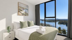 Ein Hotelzimmer mit großem Fenster, einem Bett mit grünen Kissen, einem Gemälde an der Wand und Blick auf einen See.
