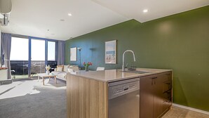 A modern kitchen with a dishwasher, a dining area with a table and chairs, and a view through large windows. - The Lakeview on Grazier (Belconnen)