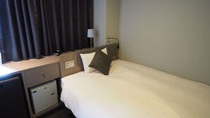 Una camera d'albergo con un letto, una scrivania con una lampada, un piccolo armadietto e una finestra con tende.