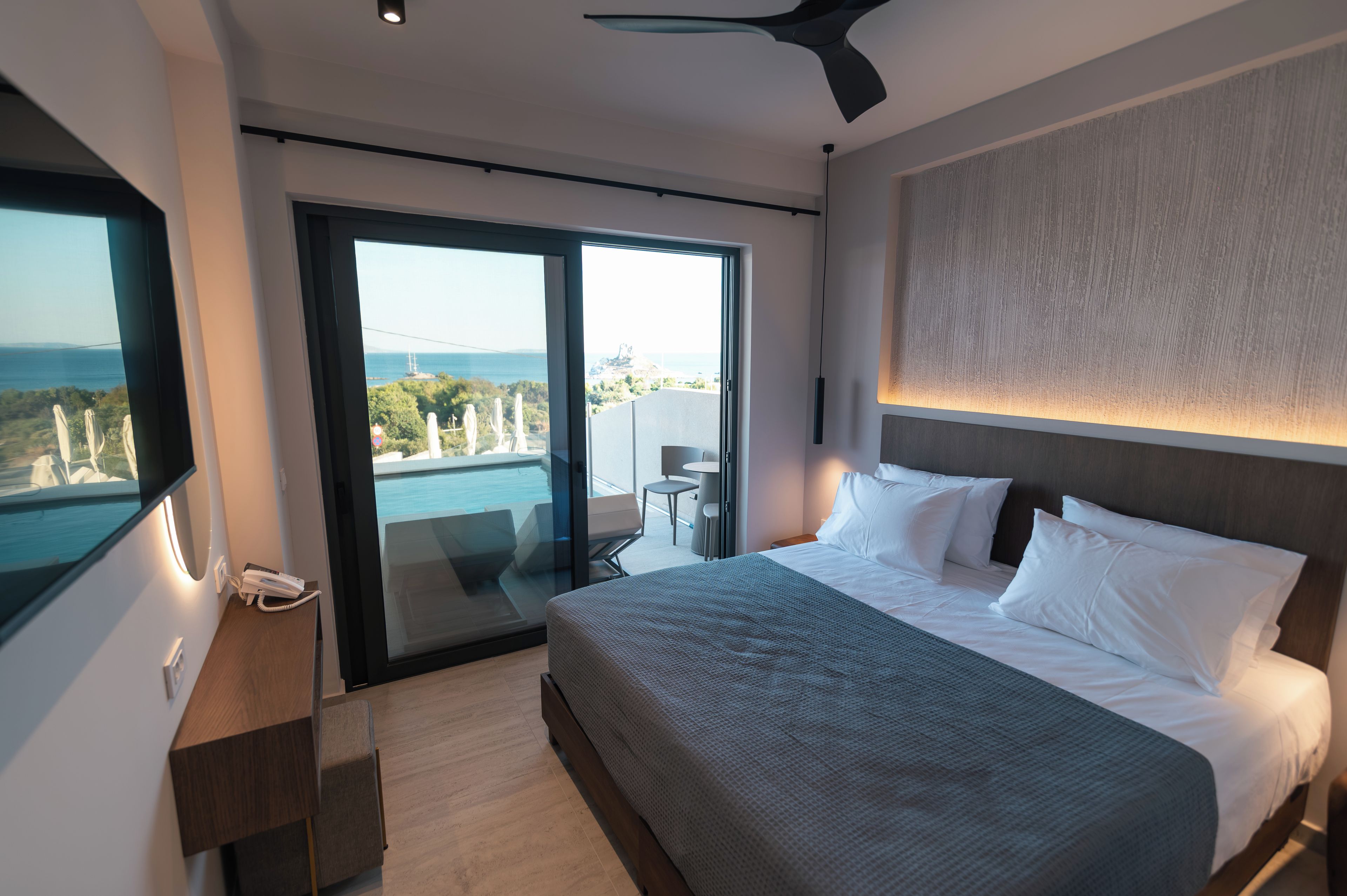 A modern hotel room with a large bed, a TV, a desk, and a balcony with a view of the sea.