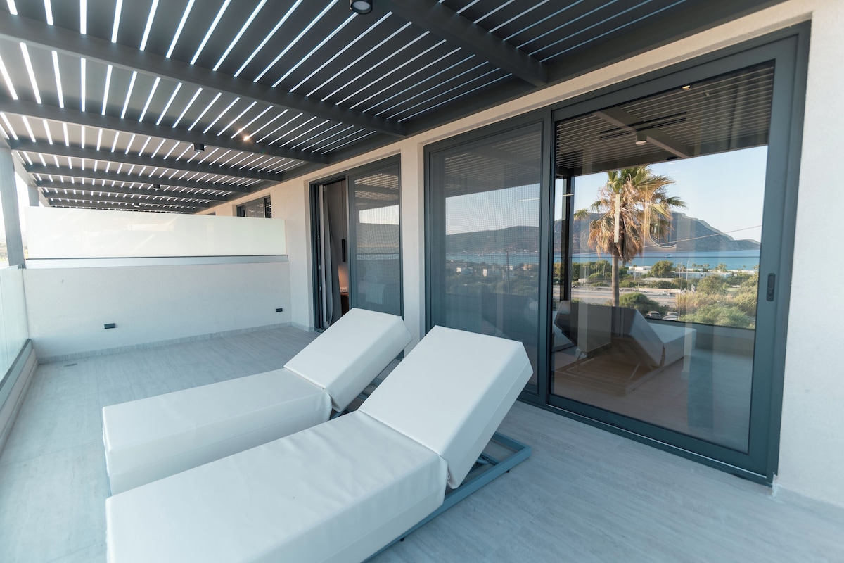 A modern balcony with two white lounge chairs, a glass door leading to a terrace, and a view of a coastal landscape.