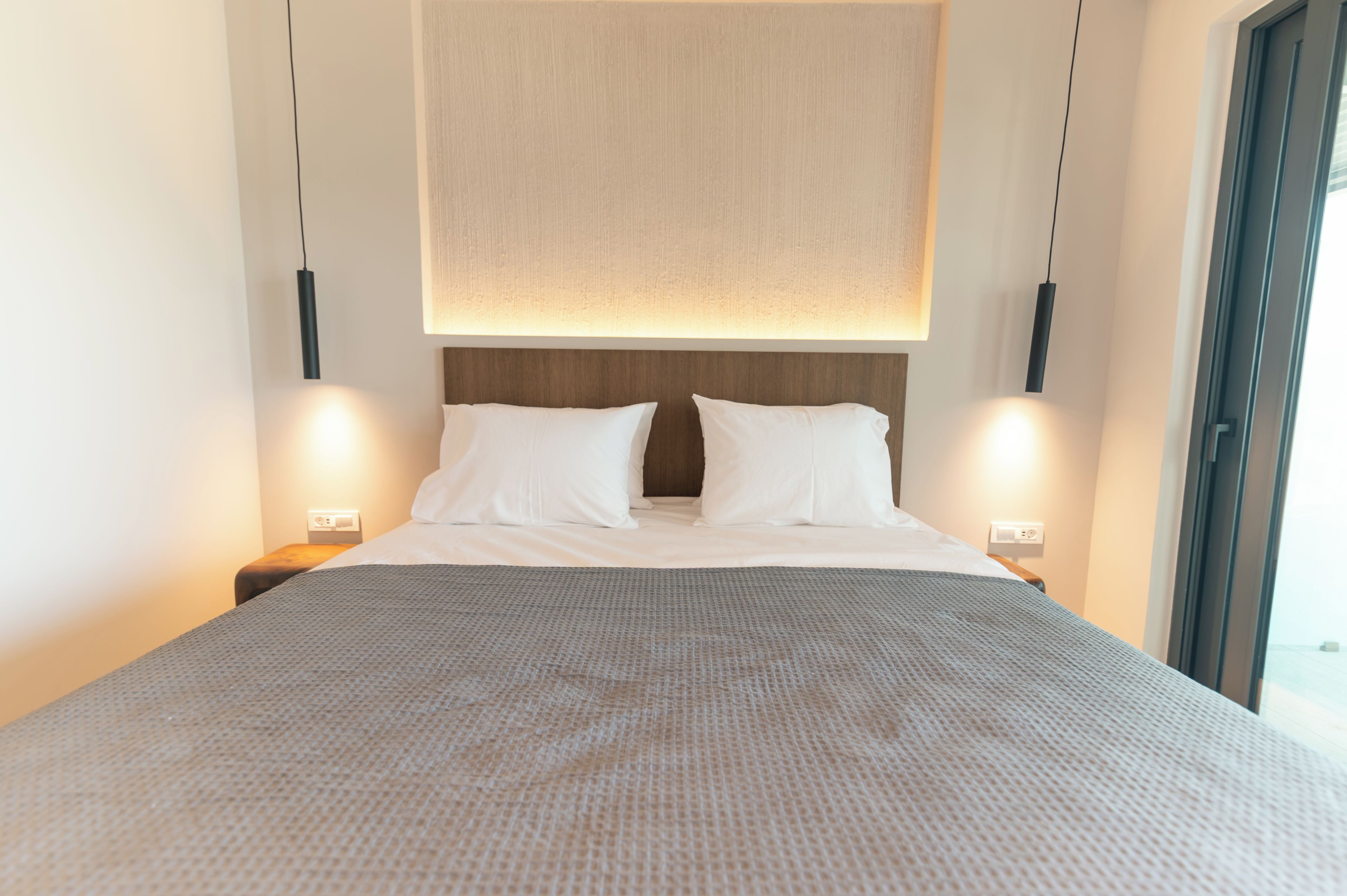 Ein ordentlich bezogenes Bett mit einer gemusterten Decke, zwei Nachttische mit Lampen und ein großes Fenster rechts.