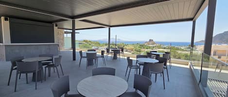 An outdoor terrace with round tables and chairs, a view of the sea and mountains, and a modern architectural design.