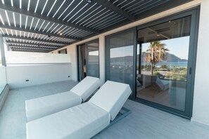 Un balcón moderno con dos sillones blancos, una puerta de cristal que da a una terraza y vistas de un paisaje costero.