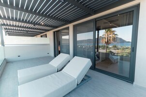 Un balcón moderno con dos sillones blancos, una puerta de cristal que da a una terraza y vistas de un paisaje costero.
