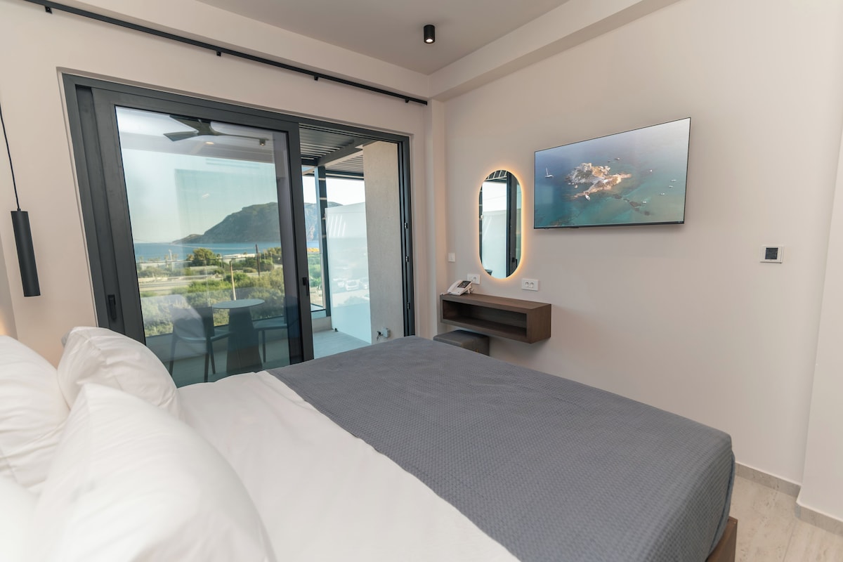 A modern hotel room with a bed, a large mirror, a flat-screen TV, and a sliding glass door leading to a balcony with a view of the sea.