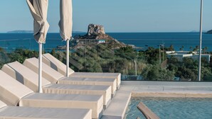 Uma área à beira da piscina com espreguiçadeiras e guarda-sóis, com vista para a paisagem costeira e uma rocha imponente.
