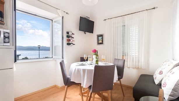 A dining area with a table set for two, a sofa, a TV mounted on the wall, and a window with a view of the sea. - Apartments Lotea - Two-bedroom Apartment With Terrace and Sea View (Konavle)