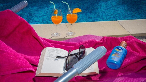 En avslappnad miljö vid poolen med ett rosa handduk, en bok, solglasögon, en flaska solskyddsmedel och två glas apelsinjuice.