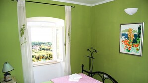 Un dormitorio con cama, ventana con cortinas, lámpara de noche, luz empotrada en la pared y un cuadro.