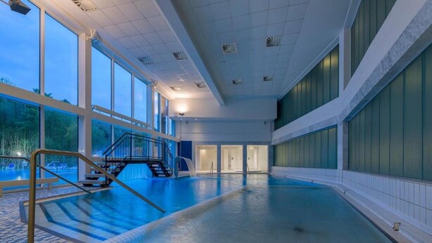 En stor inomhuspool med ett kaklat golv, en trappa som leder upp till en utsiktsplattform och stora fönster på ena sidan.