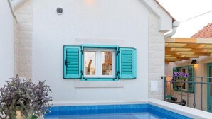 Una piscina con borde de azulejos, una ventana con contraventanas y una planta en maceta.