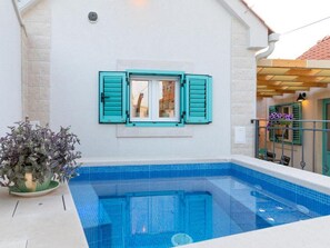 Ein Schwimmbecken mit gefliestem Rand, ein Fenster mit Rollläden und ein Blumentopf.