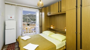 Een hotelkamer met een bed, nachtkastje, lamp, raam met uitzicht op een brug en bergen, en een kleine koelkast.