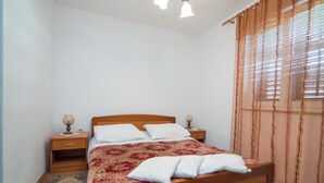 Una camera da letto con un letto di legno, cuscini bianchi, una coperta beige e rossa, due comodini con lampade, una finestra con tende e un tappeto decorato.