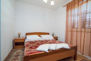 A bedroom with a wooden bed, white pillows, a red and beige bedspread, two bedside tables with lamps, a window with curtains, and a patterned rug. - Apartments Longo - Standard One Bedroom Apartment With Terrace, A3 - (Supetar)