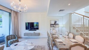 A modern dining room with a long table set for six, a television above the seating area, and a staircase to the right. - Maple Residences - Dubai Hills Estate (Dubai)