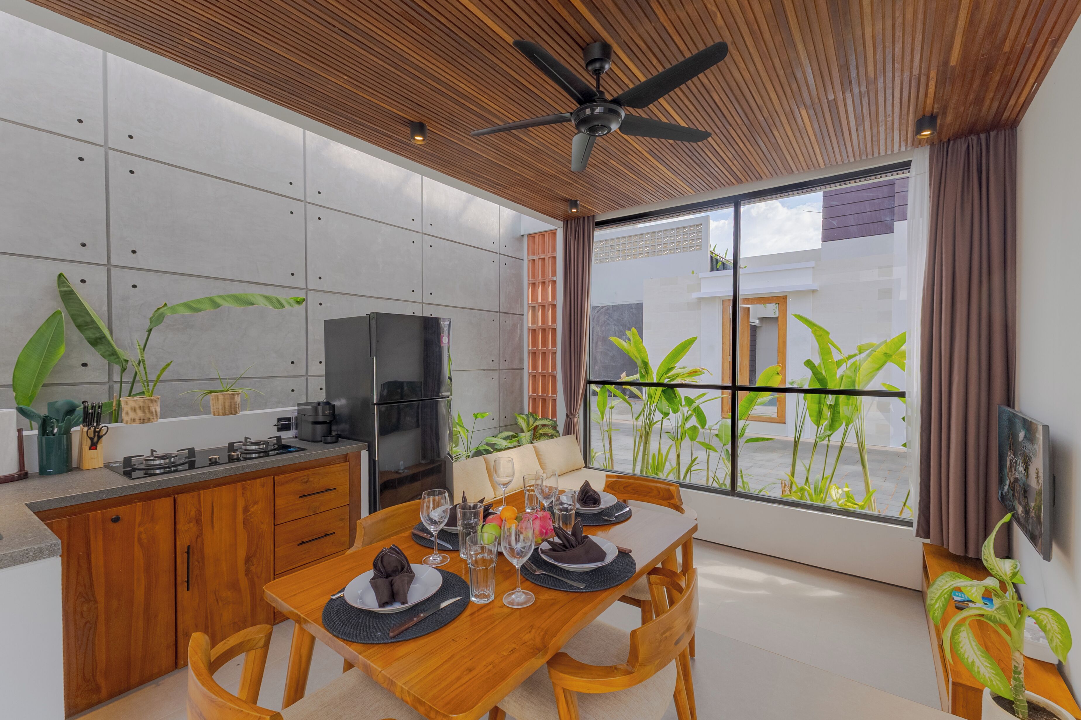 A modern kitchen with a dining area, wooden ceiling, and large windows with plants.