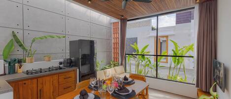 A modern kitchen with a dining area, wooden ceiling, and large windows with plants.