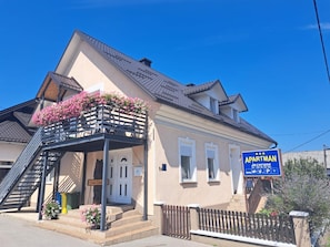 Ein zweistöckiges Gebäude mit einem Balkon, der mit rosa Blumen geschmückt ist, einer sichtbaren Treppe und einem Schild, das es als Wohnung kennzeichnet.