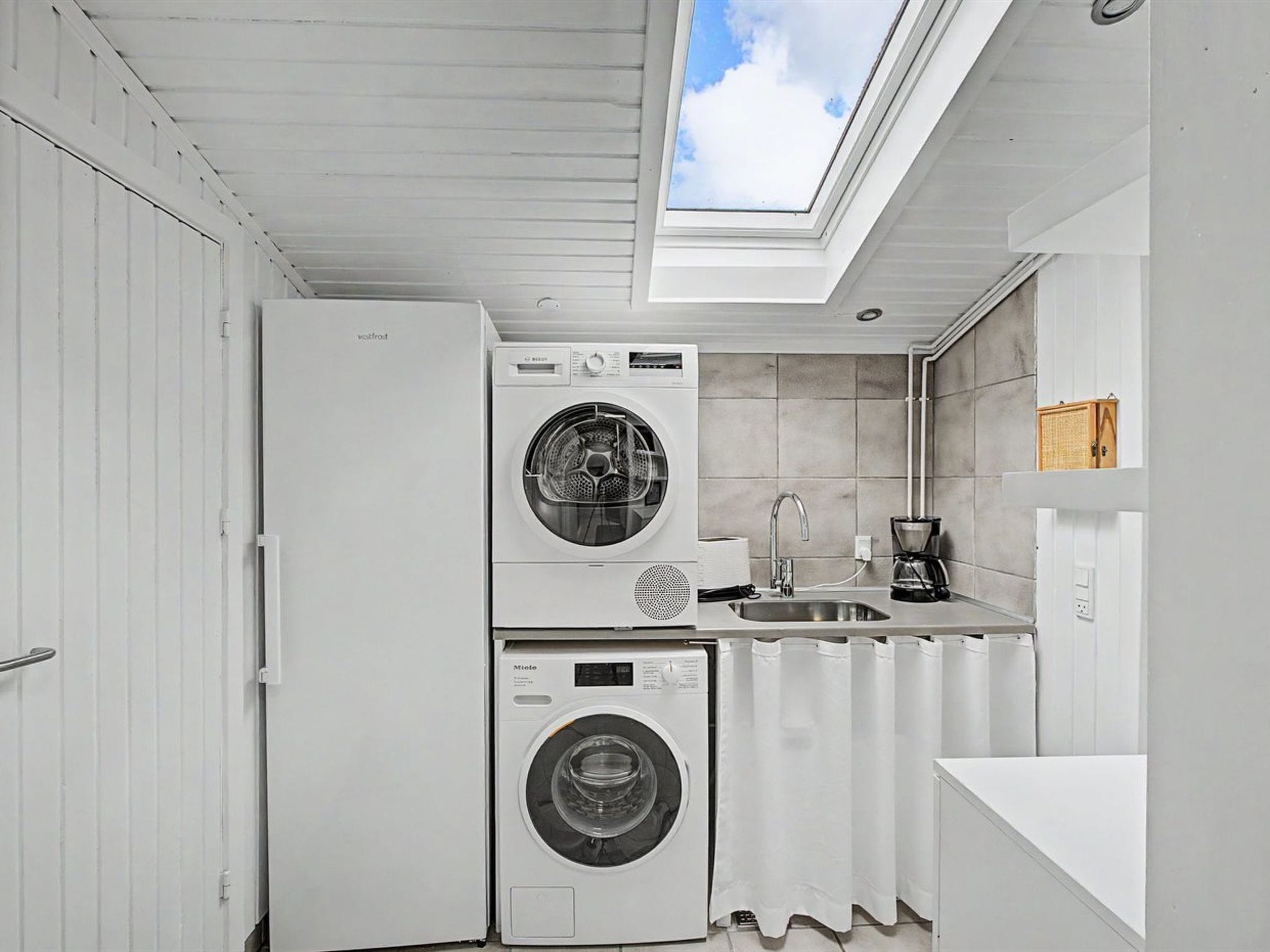 A compact laundry room with a skylight, a washing machine, a dryer, a sink, and a coffee maker.