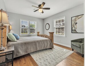 A bedroom with a bed, a ceiling fan, two windows with blinds, a wall-mounted mirror, a framed picture, and a green upholstered chair. - Watership Down: 45 Seconds from Texas A&M Campus (College Station)