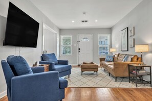A living room with a flat-screen TV mounted on the wall, two blue armchairs, a brown leather sofa with patterned cushions, a small wooden side table, and a floor lamp. - Watership Down: 45 Seconds from Texas A&M Campus (College Station)