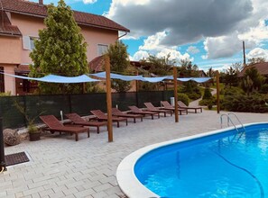 Ein Swimmingpool mit Liegestühlen und Sonnenschirmen, umgeben von einer Pflasterterrasse und einem Zaun.
