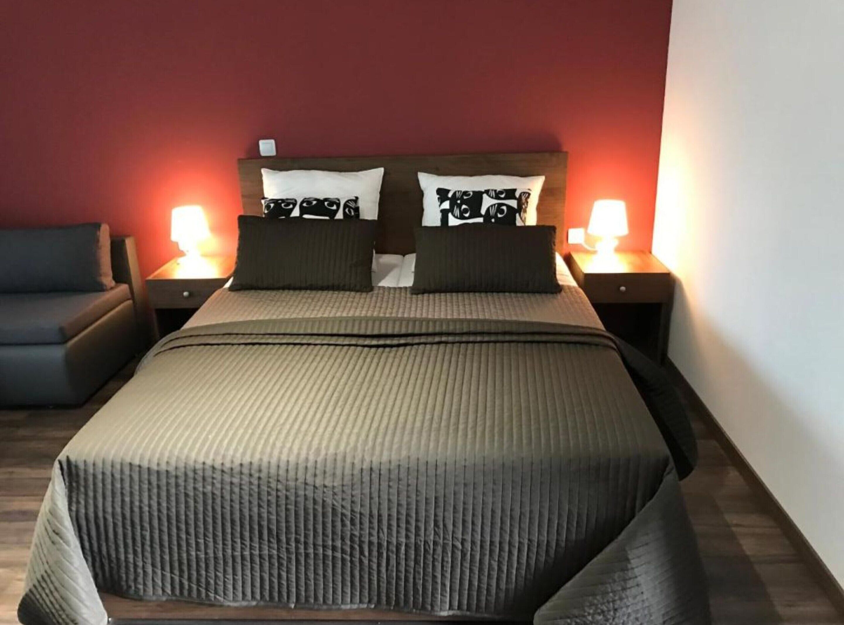 Una cama con cabecera, dos mesitas de noche con lámparas, un sofá gris y una pared roja.