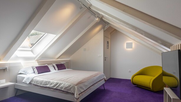 A modern bedroom with a sloped ceiling, a bed with white bedding and purple pillows, a yellow chair, and a skylight. - Rooms Lejletul - Double Room (Dubrovnik)