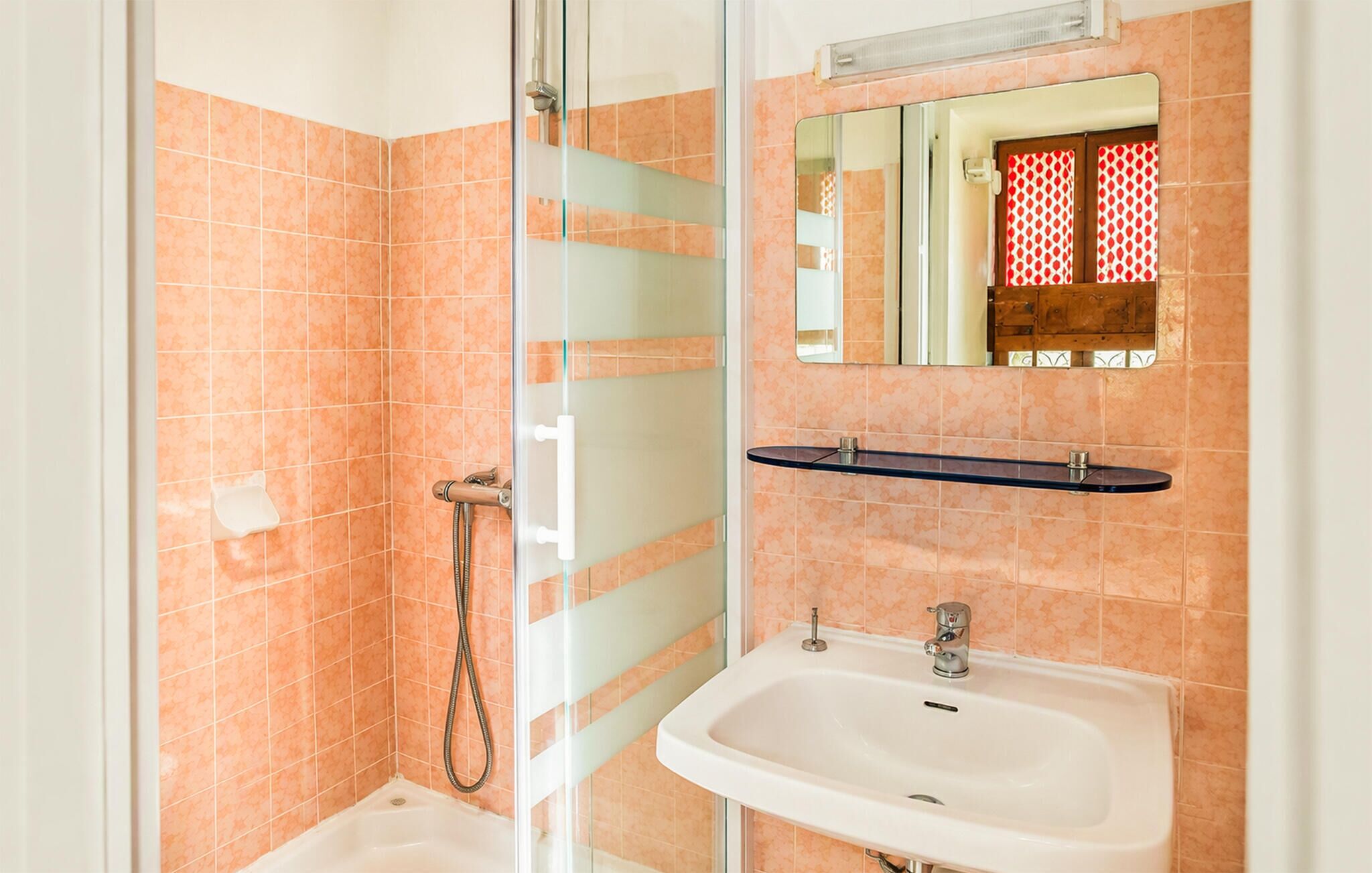 Une salle de bain avec une douche à parois de verre, un lavabo et un miroir agrémenté d’un rideau aux motifs rouges et blancs.