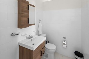 A bathroom with a white sink, wooden vanity, a toilet, and a trash bin. - Casa Magnolia Apt 6 | Santurce Multifamily House (San Juan de Puerto Rico)