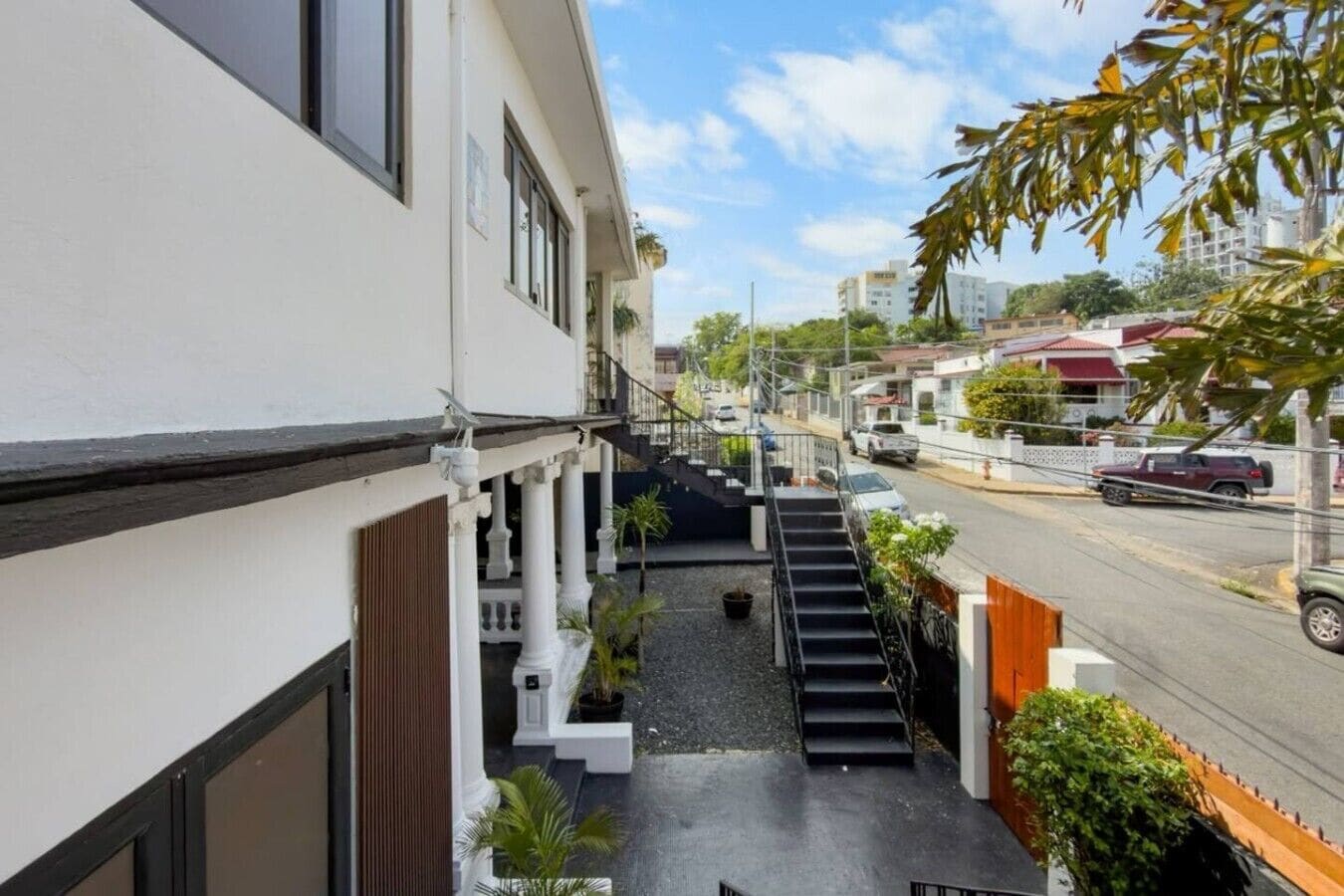 A residential building with a staircase leading to a balcony, surrounded by greenery and a street view.