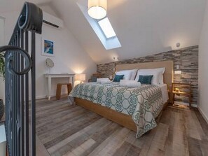 Ein Schlafzimmer mit schrägen Wänden, einem Bett mit gemusterten Textilien, einem Schreibtisch und einer Wandlampe.