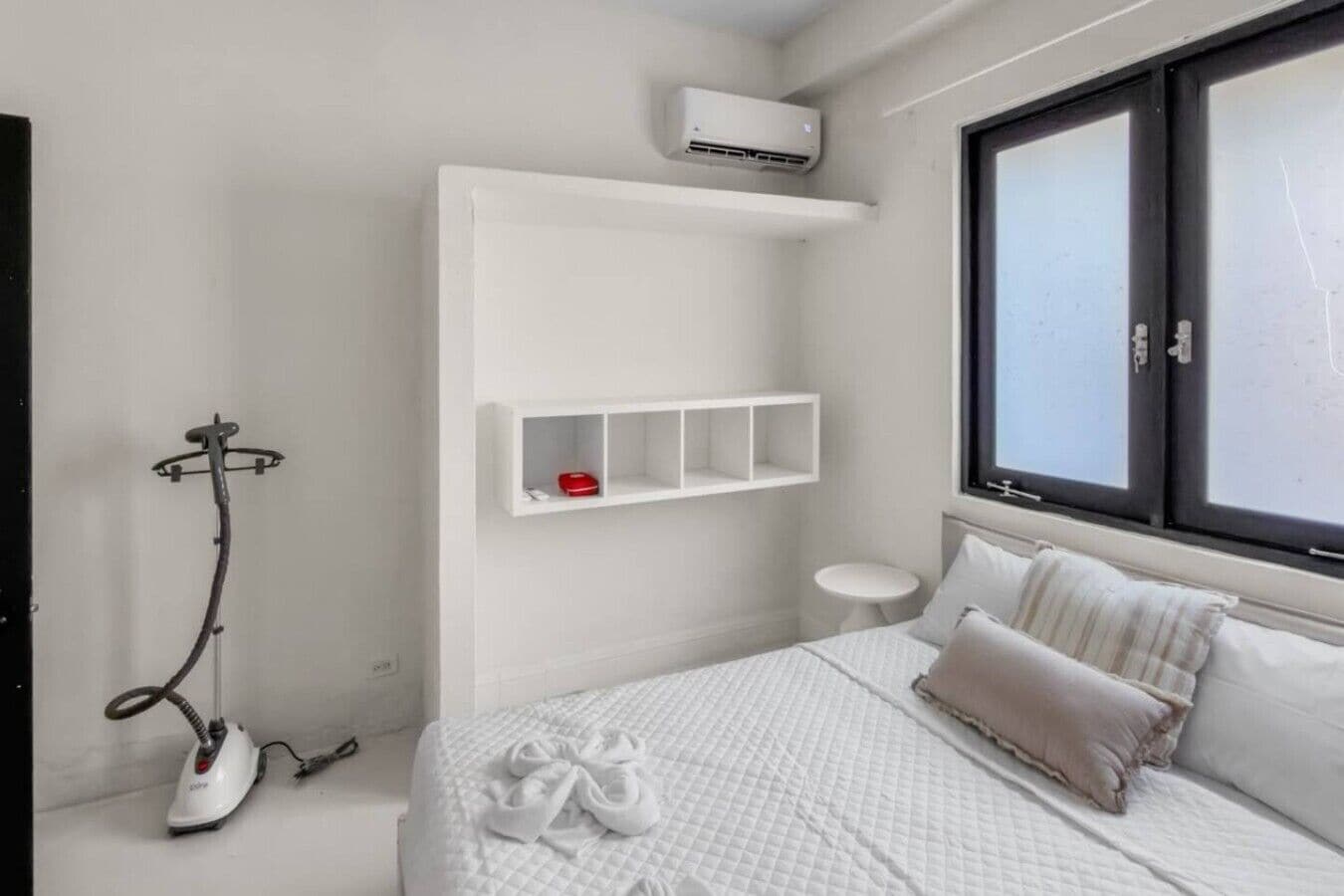 Ein Schlafzimmer mit einem Bett, einem kleinen Nachttisch, einem Fenster und einer Klimaanlage.