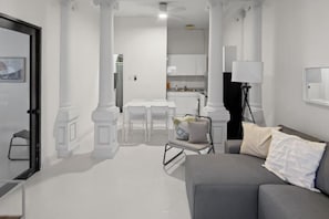 A modern living room with a grey sofa, a small table, and a chair. There is a kitchen area in the background with white cabinets and a countertop. - Casa Magnolia Apt 1 | Santurce Multi Family House (San Juan de Puerto Rico)