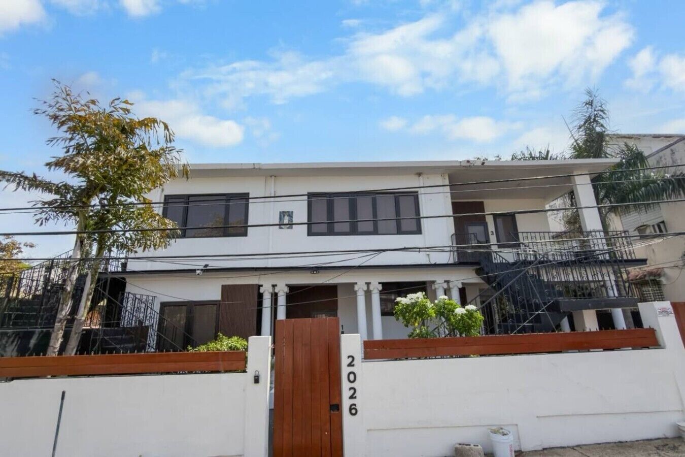 Um edifício residencial de dois andares com fachada branca, um portão de madeira e o número '2026' visível no portão.