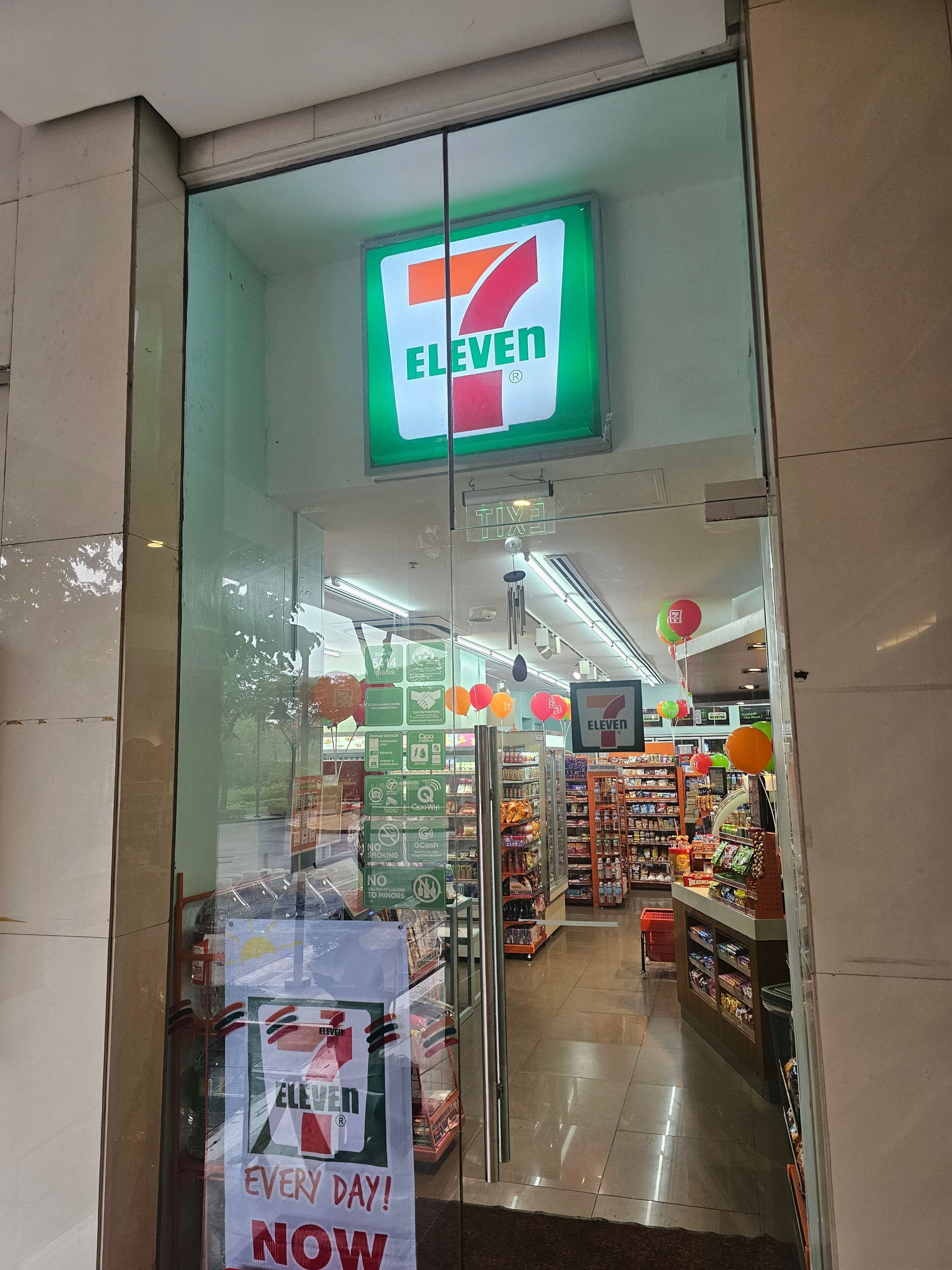 Entrada a una tienda 7-Eleven con puerta de vidrio, un letrero sobre la puerta y un anuncio en el vidrio.