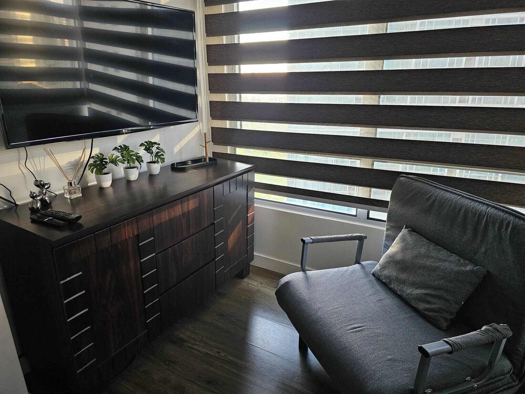 A modern living room with a dark wood cabinet, a flat-screen TV, a grey sofa, and a window with horizontal blinds.