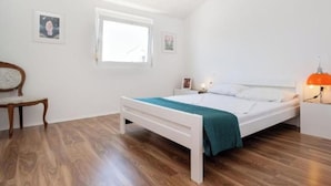 Un dormitorio con cama, una silla, una mesita de noche, una lámpara y una ventana.