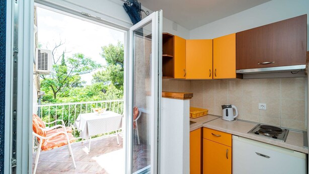 A modern kitchen with orange cabinets, a white countertop, and a view of a balcony with outdoor furniture. - Apartment & Rooms Papa - Studio Apartment With Balcony and Partial Sea View (Zupa dubrovacka)