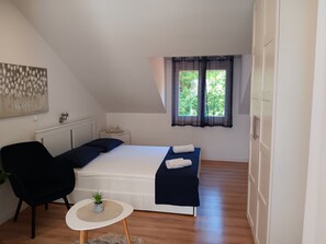 Ein Schlafzimmer mit einem Bett, einem Sessel, einem kleinen Tisch mit Pflanze, einem Fenster mit Vorhängen und einer Heizung.