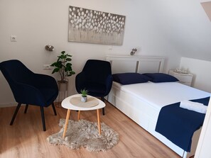Ein Schlafzimmer mit einem Bett, zwei Stühlen, einem kleinen Tisch, einer Zimmerpflanze und einem Bild an der Wand.