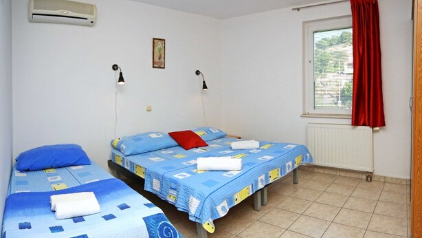 A room with two beds, each with a blue and white patterned bedspread, a red pillow, and white towels. There are two wall-mounted lamps, a window with red curtains, and a radiator. - Apartments & Rooms Villa Petra - Double Room (Sibenik)