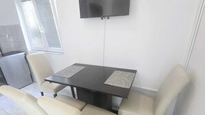 A dining area with a table and four chairs, a television mounted on the wall, and a window with blinds. - Apartments Mateo - One Bedroom Apartment - White (Vodice)