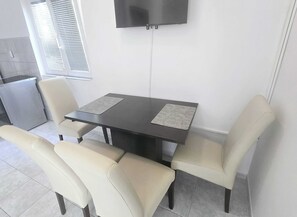 A dining area with a table and four chairs, a television mounted on the wall, and a window with blinds. - Apartments Mateo - One Bedroom Apartment - White (Vodice)