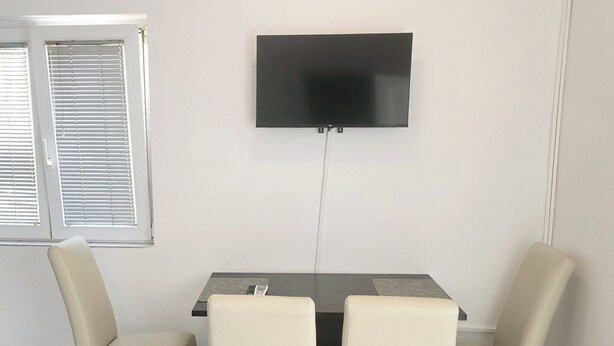 A dining area with a table and two chairs, a television mounted on the wall, and a window with blinds. - Apartments Mateo - One Bedroom Apartment - White (Vodice)