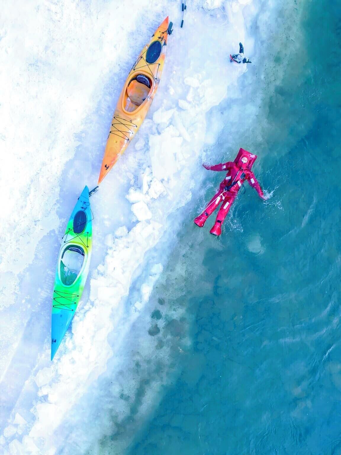 A kayaker in a red wetsuit and two kayaks on a frozen body of water.